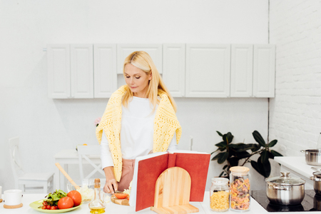 beautiful blonde woman cooking salad with cookbook at kitchenの写真素材