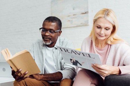 african american man in glasses reading book while mature woman reading travel newspaper