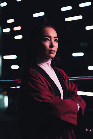 attractive asian girl in burgundy kimono standing with crossed arms on street with neon light in evening, city of future conceptの写真素材