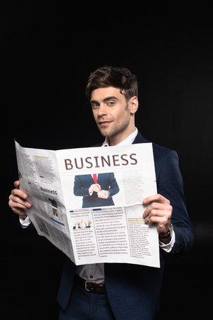 attractive young businessman with newspaper looking at camera isolated on blackの写真素材