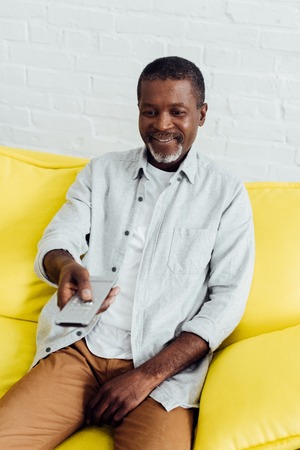 smiling handsome african american man sitting on sofa with tv remote controller