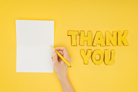 cropped person holding pen above blank sheet and thank you lettering in cookies isolated on yellow backgroundの写真素材