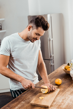 smiling young man in pajamas cutting lemon on chopping board in kitchenの写真素材