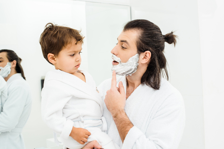 Father showing toddler son shaving foam on faceの写真素材