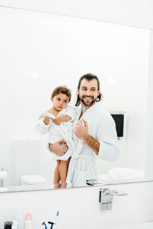 Dad and toddler son looking in mirror at home in white bathroom