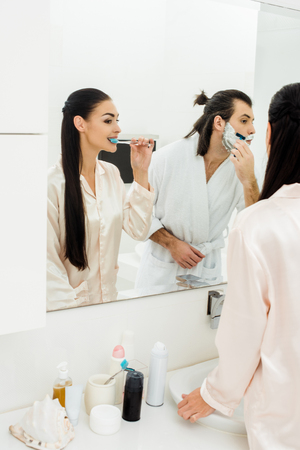 beautiful woman brushing teeth and handsome man shaving in front of mirror in bathroomの写真素材