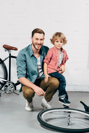 happy man sitting with little son and looking at camera in bicycle workshopの写真素材