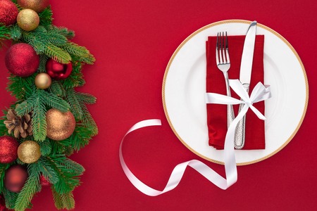 top view of fork and knife wrapped by festive ribbon on plate near evergreen tree branches with christmas balls isolated on redの写真素材