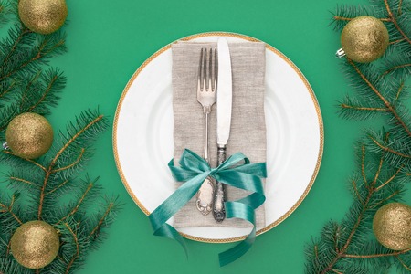view from above of fork and knife wrapped by festive ribbon on plate surrounded by evergreen tree branches with christmas balls isolated on greenの写真素材