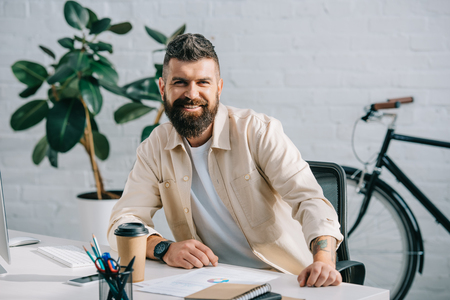 Bearded businessman sitting in modern office and smiling at cameraの写真素材