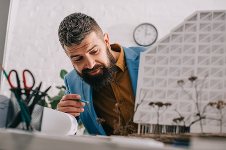 focused adult male architect in formal wear pointing at house model and working on projectの写真素材