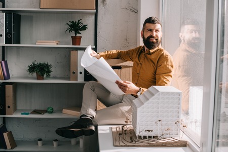 smiling bearded adult male architect sitting near house model, holding blueprint and working on project in officeの写真素材