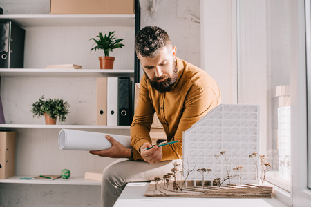 focused bearded adult male architect holding blueprint, pointing at house model and working on project in officeの写真素材