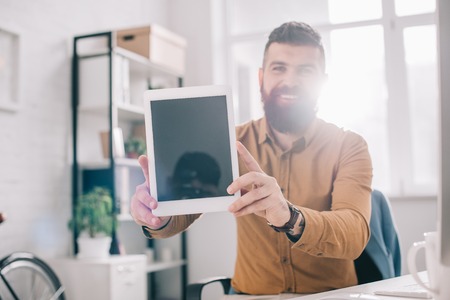 smiling adult business man presenting digital tablet with blank screen at workplace with backlit