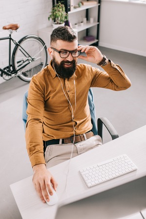 smiling bearded adult businessman in earphones sitting and working at computer desk in officeの写真素材