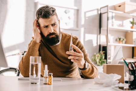 sick businessman holding medical thermometer and sitting at office desk with glass of water and...