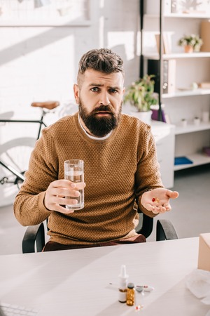 sick businessman sitting at office desk, looking at camera, holding glass of water and medicine in...