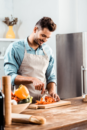 smiling young man in apron chopping fresh vegetables in kitchenの写真素材