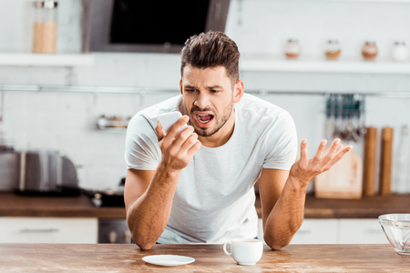 emotional young man yelling at smartphone while drinking coffee in kitchenの写真素材