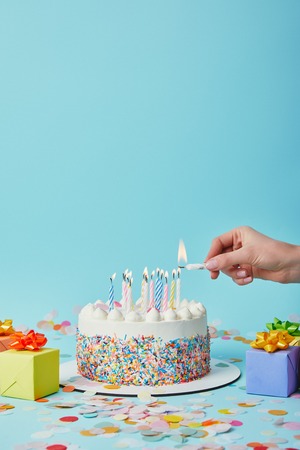 Partial view of woman lighting birthday cake on blue background with gifts and confettiの写真素材