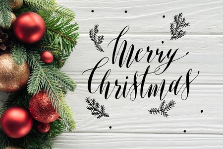 top view of wreath with red christmas toys on white wooden surface with merry christmas lettering