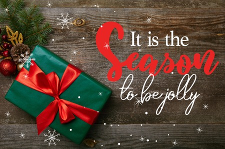 top view of wrapped green present with red ribbon and christmas toys on wooden background with "it is the season to be jolly" inspirationの写真素材