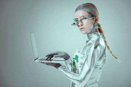 side view of silver robot holding laptop and looking at camera isolated on grey, future technology conceptの写真素材