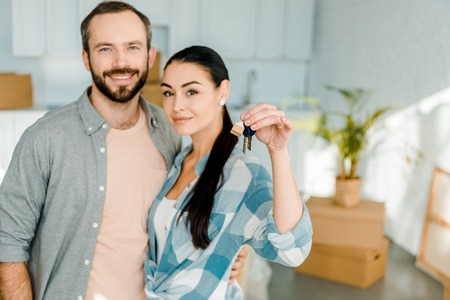 happy couple looking at camera and wife holding keys from new house, moving concept