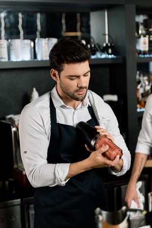 handsome barman in apron preparing cocktail in glass with shakerの写真素材