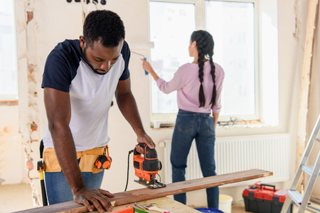 handsome african american man working with jigsaw while his girlfriend working behind during renovation at homeの写真素材