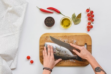 cropped image of tattooed woman cutting fish by knife on wooden board near ingredients on tableの写真素材