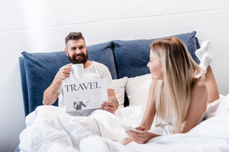 Smiling bearded man holding coffee cup and reading travel newspaper while young girl using smartphone in white bedの写真素材
