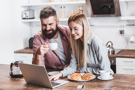 young woman using laptop at kitchen table while man holding white cupの写真素材