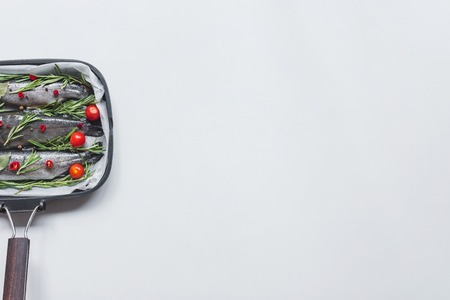 elevated view of fish with rosemary, bay leaves and cherry tomatoes in tray with baking paper on...