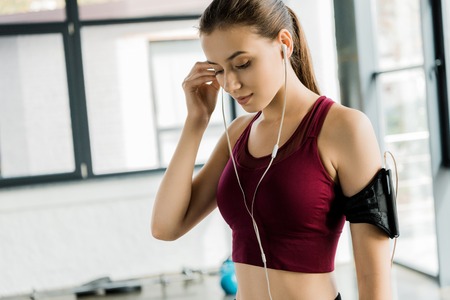 beautiful slim sportswoman in smartphone armband putting on earphones at gym