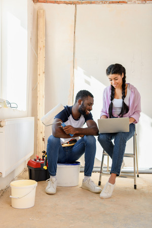 happy african american couple with laptop and paint roller during renovation of homeの写真素材
