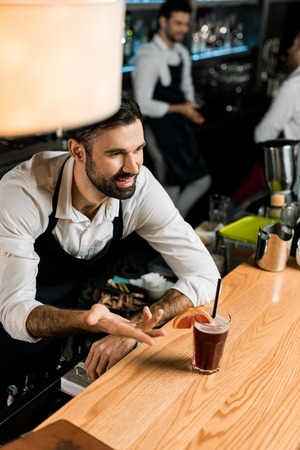 handsome smiling bartender serving cocktail with grapefruit slice and straw at wooden counter