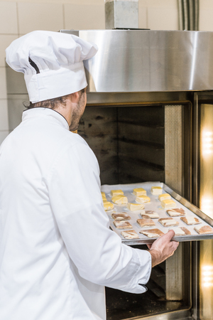 male baker in white chefs uniform putting baking tray with uncooked dough in ovenの写真素材