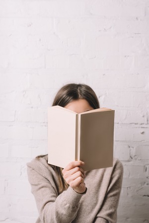 girl hiding face behind book near white brick wallの写真素材