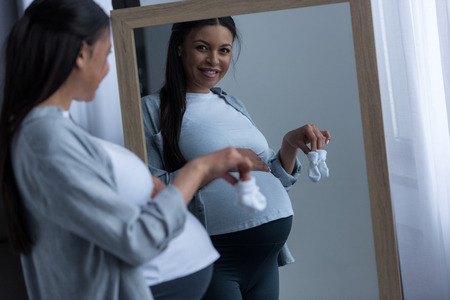 african american pregnant woman holding little socks and looking at reflection in mirrorの写真素材