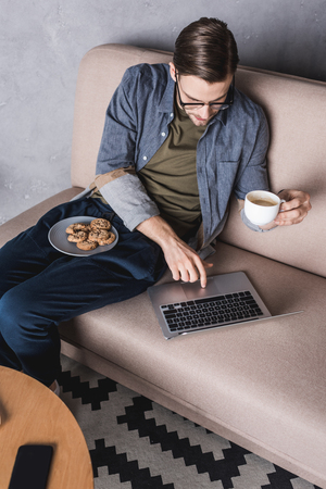 young handsome man working with laptop and drinking coffee on couchの写真素材