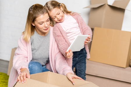 happy mother and daughter using digital tablet while packing cardboard boxes