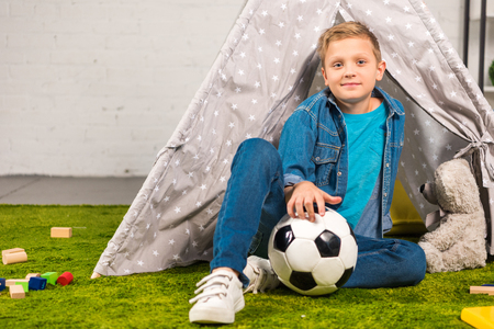 positive little boy sitting with soccer ball near tent at homeの写真素材