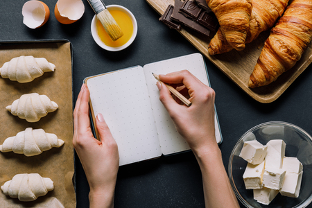 partial view of woman writing recipe in textbook at table with dough for croissants on tray and ingredientsの写真素材