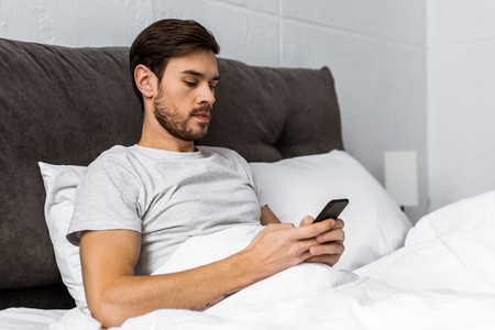 handsome bearded young man using smartphone in bed in the morningの写真素材