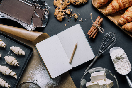 view from above of tray with dough for croissants, ingredients and blank textbook on black tableの写真素材