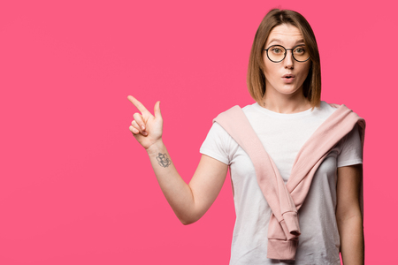 surprised young woman in eyeglasses pointing by finger isolated on pinkの写真素材