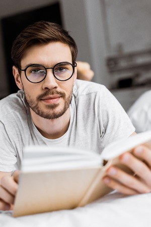 young man in eyeglasses holding book and looking at camera while lying on bedの写真素材