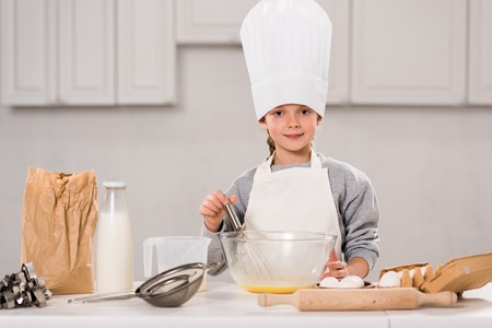 smiling child in chef hat whisking eggs in bowl at table in kitchenの写真素材