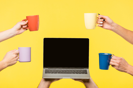 cropped shot of hands holding cups and laptop with blank screen isolated on yellowの写真素材